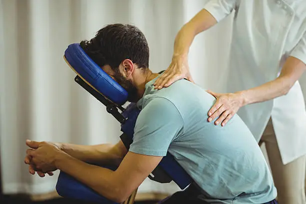 Male leaning into a massge chair with his face in the face cradle whilst a therapist uses acupressure techniques to perform seated chair massage.